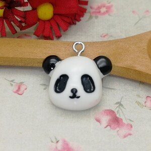 May include: A white and black panda bear charm with black eyes and a small black nose. The charm is made of polymer clay and has a silver loop for attaching to a necklace or bracelet.