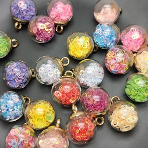 Könnte beinhalten: Sortiment an bunten Glaskugeln mit verschiedenen Konfetti im Inneren. Die Anhänger befinden sich auf einer schwarzen Oberfläche und haben goldene Ösen zum Aufhängen.