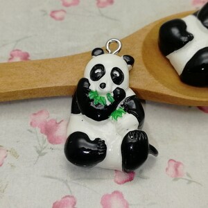 May include: Two black and white panda bear charms with green bamboo leaves. The charms are sitting on a wooden spoon. The spoon is on a white background with pink flowers.