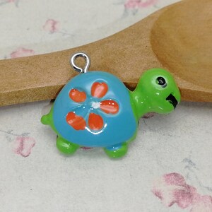 Puede incluir: Un pequeño amuleto de tortuga azul y verde con una flor naranja en su caparazón. El amuleto tiene un lazo plateado para sujetarlo a un collar o pulsera.