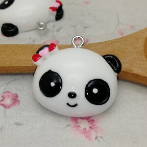 May include: Two white panda bear charms with black eyes and a black nose. One panda has a pink and red bow on its head. The charms are on a wooden spoon and a floral background.