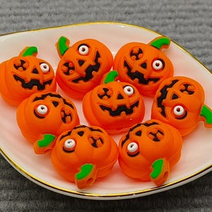 Op de afbeelding: Een wit, bladvormig bord met een gouden rand bevat zeven oranje pompoenversieringen. Elke pompoen heeft een gesneden gezicht met zwarte ogen en mond, en een groene stengel. Halloween-thema decoraties.