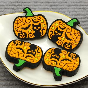 Op de afbeelding: Vier Halloween pompoen bedels met zwarte kat en vleermuis ontwerpen, gepresenteerd op een wit bord met een gouden rand. De pompoenen zijn oranje met groene stelen en zwarte contouren.