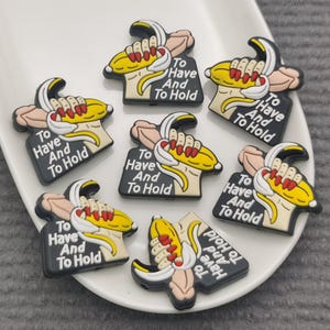 May include: A collection of shoe charms featuring a cartoon banana being held by a hand. Each charm has a black tag with the text "To Have And To Hold". The banana is yellow with red accents.