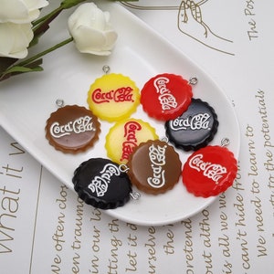 May include: Assortment of eight colourful plastic bottle cap charms with the Coca-Cola logo in white lettering. The charms are on a white plate.