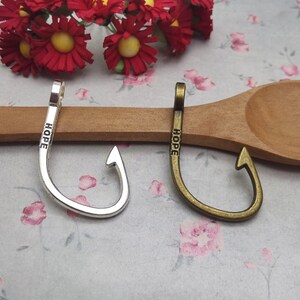 May include: Two silver and bronze fishing hooks with the word "HOPE" engraved on them. The hooks are on a wooden spoon.