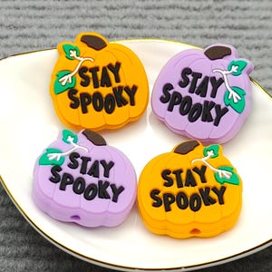 Op de afbeelding: Vier pompoenvormige bedels in oranje en paars, elk met de tekst "STAY SPOOKY". Ze liggen op een wit bord met een gouden rand. De pompoenen hebben groene bladeren en bruine stelen, perfect voor Halloween.