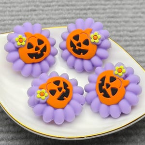 Op de afbeelding: Vier Halloween-thema decoratieve versieringen. Elk heeft een lachend pompoengezicht in oranje, geplaatst op een paarse bloem met gele accenten. De items zijn gerangschikt op een wit, bladvormig schaaltje met een gouden rand.