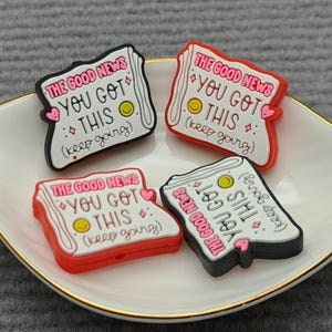 Puede incluir: Cuatro colgantes rectangulares coloridos con el texto "THE GOOD NEWS YOU GOT THIS (keep going)" se muestran en un plato blanco en forma de hoja con borde dorado. Los colgantes son negros, rojos y blancos.