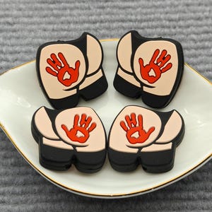 May include: Four novelty butt-shaped charms with a red handprint design, displayed on a white, gold-rimmed dish. The charms are tan, black, and red, and are arranged in a square formation.