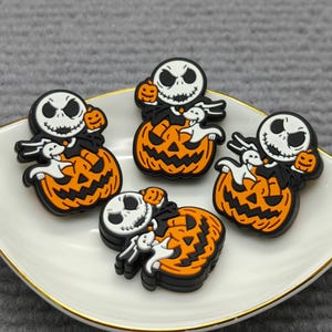 Op de afbeelding: Vier Halloween-schoenbedels met Jack Skellington-personages met een pompoen- en spookontwerp. De bedels zijn zwart, wit en oranje en zijn gerangschikt op een wit bord met een gouden rand. Het bord staat op een grijze ondergrond.