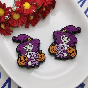 Op de afbeelding: Twee Halloween-charms met een heks met paars haar en een puntmuts, die een paarse kat vasthoudt en geflankeerd wordt door twee oranje pompoenen. De bedels liggen op een wit bord met rode bloemen.