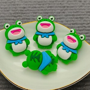 May include: Four green frog figurines with white bellies, blue bow ties, and pink mouths rest on a white plate with a gold rim. One frog is lying down. The plate is on a gray surface.