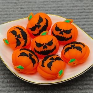 Op de afbeelding: Een wit en goudomrand bordje bevat zeven oranje pompoenvormige kralen. Elke kraal heeft een gesneden zwart gezicht en een groene stengel. De kralen zijn een Halloween-thema knutselbenodigdheid.