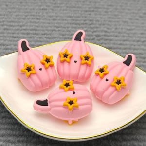 Op de afbeelding: Vier roze pompoenvormige decoraties met zwarte stelen en gele steraccenten zijn gerangschikt op een wit, bladvormig bord met een gouden rand. De pompoenen hebben kleine zwarte stippen als ogen.