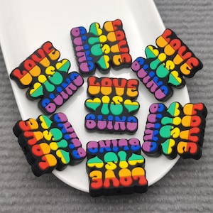 May include: A collection of colorful rubber charms with the phrase "LOVE IS BLIND" in rainbow colors and black outlines. The charms are arranged on a white, oval-shaped plate.