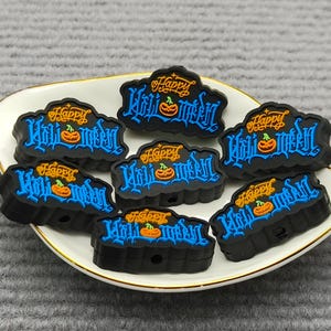 Op de afbeelding: Een verzameling zwarte Halloween-bedels met de tekst "Happy Halloween" in blauwe letters, elk met een oranje pompoen. De bedels liggen op een wit bord met een gouden rand.