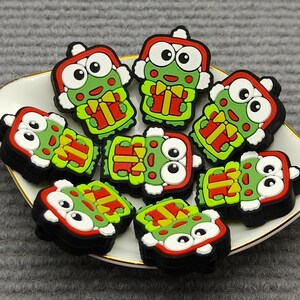 May include: A collection of cartoon frog-shaped charms in green, red, and white, arranged on a white and gold-rimmed dish. The charms have a gift box design and are set against a gray background.