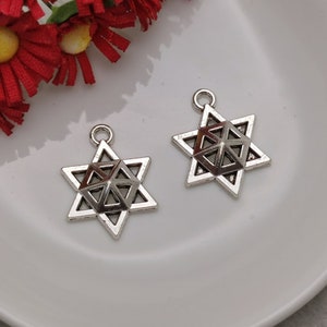 May include: Two silver-toned metal charms in the shape of a six-pointed star with a geometric design inside. The charms are on a white surface.