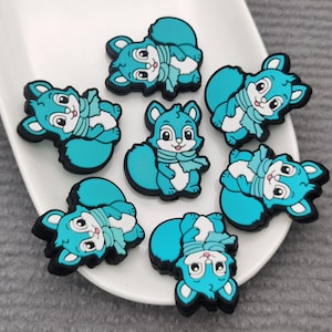 May include: A collection of seven teal squirrel charms with black outlines and white accents, each wearing a scarf. The charms are arranged on a white, oval-shaped dish, set against a gray background.