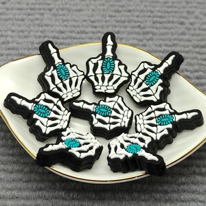 以下が含まれることがあります： 白と黒の骸骨の手のチャームのコレクション。中央はターコイズ色で、金色の縁取りのある白い皿の上に配置されています。チャームは中指のジェスチャーの形をしています。