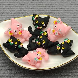 Peut inclure: Une collection d'ornements en forme de chat noirs et roses, ornés de motifs floraux, disposés sur une assiette blanche bordée d'or. Les chats ont des yeux jaunes et des moustaches blanches. L'assiette est posée sur un fond gris texturé.