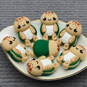May include: A white, leaf-shaped dish with a gold rim holds several turtle-shaped figurines. The turtles have tan heads with brown spots, white bellies, and green and tan shells. One turtle is flipped over, revealing a green shell.