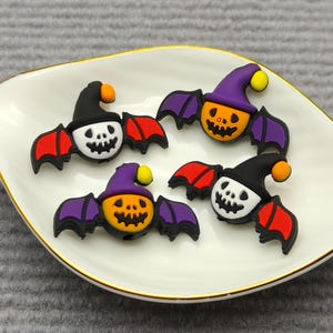Op de afbeelding: Vier Halloween-vleermuisdecoraties op een wit, bladvormig schaaltje met een gouden rand. De vleermuizen hebben oranje pompoengezichten, paarse vleugels en dragen heksenhoeden in zwart of paars. De vleugels zijn rood.