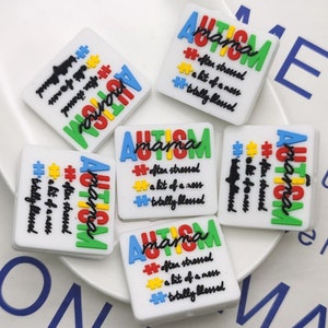Könnte beinhalten: Fünf bunte quadratische Silikon-Anhänger mit dem Text "AUTISM mama" und dem Satz "oft gestresst, ein bisschen Chaos, total gesegnet" in schwarzer Schrift. Die Anhänger sind mit Puzzle-Teilen in Rot, Gelb, Blau und Grün verziert.