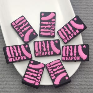 May include: A collection of black rectangular patches with pink illustrations of sex toys and the word "WEAPON" in white. The patches are arranged on a white plate.