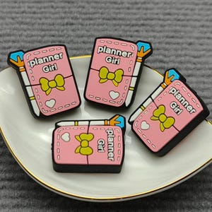 May include: Four pink planner charms with the text "planner Girl" and a yellow bow design, arranged on a white and gold-rimmed dish. The charms have a black border and a small heart detail. The background is a gray surface.