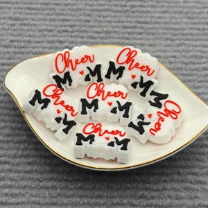 Puede incluir: Un conjunto de dijes blancos y rojos de "Cheer Mom" sobre un plato blanco con borde dorado. Los dijes tienen forma de nube con la palabra "Cheer" en rojo y "MOM" en negro.