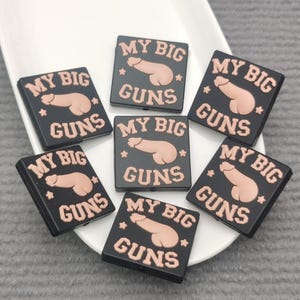 May include: Seven black square charms with the text "MY BIG GUNS" in light pink. Each charm features a graphic of a penis. The charms are arranged on a white plate.