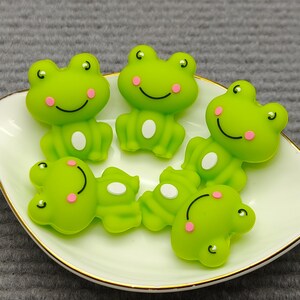 May include: A collection of six green frog-shaped embellishments with white bellies and pink cheeks, arranged on a white, leaf-shaped dish with a gold rim. The frogs have smiling faces.