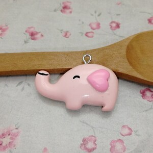 May include: A pink elephant charm with a heart-shaped ear and black eyes. The charm is made of polymer clay and has a silver loop for attaching to a necklace or bracelet.