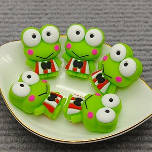 May include: A collection of six green frog figurines with large white eyes and pink cheeks, arranged on a white, leaf-shaped dish with a gold rim. The frogs wear red and white striped outfits.