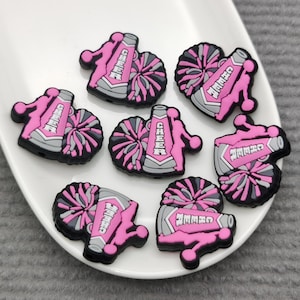 Puede incluir: Siete abalorios de megáfono de animadora rosa y gris con pompones. Los megáfonos tienen las palabras "Cheer" y "Go Team" escritas en ellos.