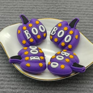 Op de afbeelding: Vier paarse pompoenvormige decoraties met het woord "BOO" in het wit, geaccentueerd met gele sterren. De decoraties liggen op een wit bord met een gouden rand, perfect voor Halloween.
