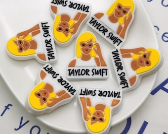 May include: Seven white and yellow plastic charms with the text "Taylor Swift" on each charm. The charms feature a cartoon image of a woman with blonde hair and sunglasses.