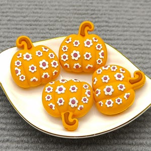 Op de afbeelding: Vier oranje pompoenvormige decoraties met witte en paarse bloemaccenten liggen op een wit, bladvormig bord met een gouden rand. De pompoenen hebben gekrulde stelen.