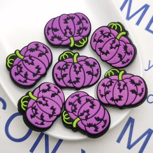 Op de afbeelding: Zeven paarse en groene Halloween-pompoencharms met vleermuis-silhouetten. De charms zijn gemaakt van rubber en hebben een diameter van ongeveer 5 cm.
