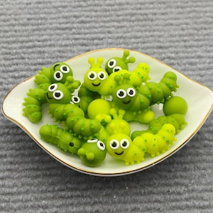 Könnte beinhalten: Eine Ansammlung kleiner, hellgrüner raupenförmiger Anhänger mit weiß-schwarzen Augen und lächelnden Gesichtern. Diese Miniaturfiguren liegen zusammen in einer weißen, blattförmigen Keramikschale mit einem dünnen, goldfarbenen Rand.