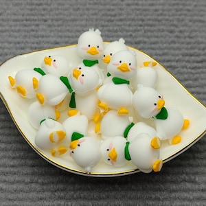 May include: A collection of small, white duck figurines with yellow beaks and feet, and green scarves, arranged in a white, leaf-shaped dish with a gold rim. The ducks are clustered together, creating a playful and whimsical scene.