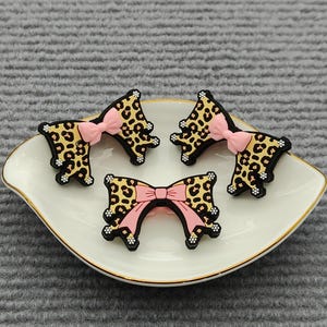 Op de afbeelding: Vier decoratieve strikvormige versieringen met een luipaardprintontwerp en roze strikken, gerangschikt op een wit, geschulpte schaal met een gouden rand. De strikken hebben zwarte contouren.