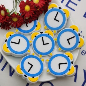 Puede incluir: Una colección de adornos con forma de reloj con esferas blancas, bordes azules, detalles amarillos y manecillas negras. Los relojes están dispuestos sobre un plato blanco con flores rojas. La imagen también contiene el texto "E LIT" y "SON of yo".