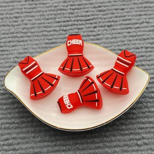 Puede incluir: Cuatro dijes de falda de animadora rojos con detalles de rayas blancas y negras. Dos de los dijes presentan la palabra CHEER en letras blancas. Estos pequeños artículos moldeados están dispuestos en un plato blanco con un borde dorado.
