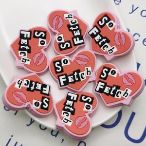 Puede incluir: Siete encantos en forma de corazón rosa con el texto "So Fetch" en letras negras. Los encantos tienen una impresión de labios rojos.