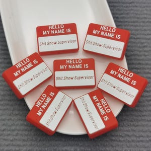 May include: Red and white name tags with the text "HELLO MY NAME IS" and "Shit Show Supervisor". The name tags are arranged on a white plate, set against a gray background. The name tags are a novelty item.