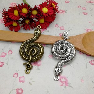 May include: Two silver snake charms, one with a bronze finish, coiled in a spiral shape. The charms are on a wooden surface with a spoon.