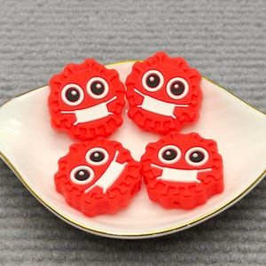 Puede incluir: Cuatro figuras rojas, tipo caricatura, con mascarillas blancas y ojos grandes, dispuestas sobre un plato blanco en forma de hoja con borde dorado. Las figuras tienen un diseño circular texturizado.